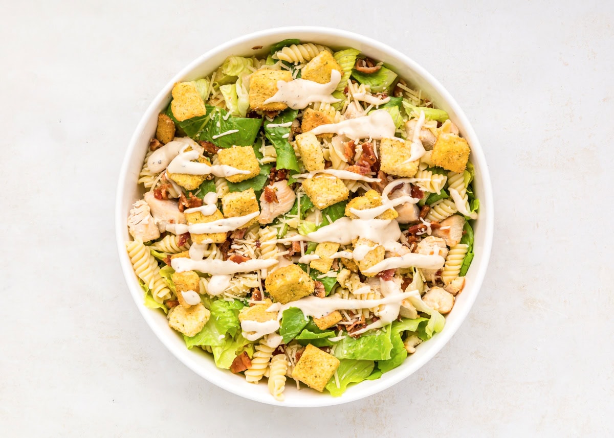 Dressing drizzled on top of the past and lettuce mixture in a white bowl.