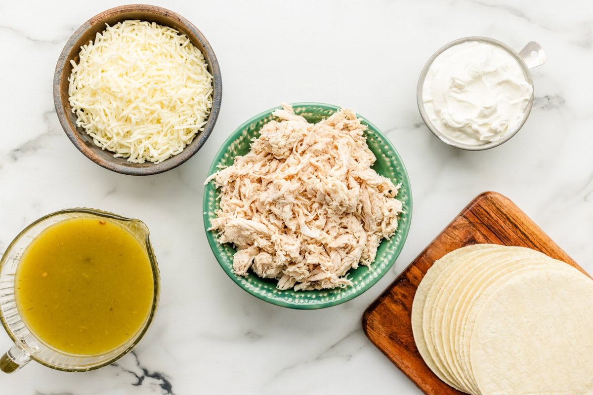 Shredded chicken, green enchilada sauce, cheese, tortillas, and sour cream on a kitchen counter.