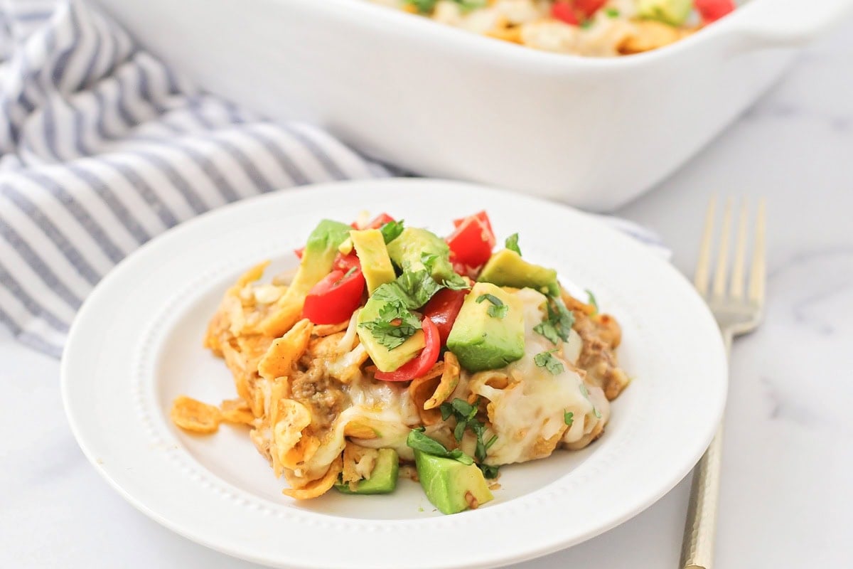 A scoop of Frito pie recipe on a white plate topped with chopped tomatoes and avocados.