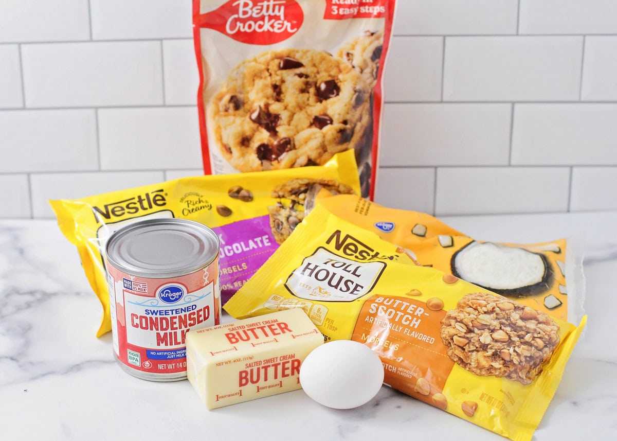 Our favorite magic cookie bars are the ultimate easy dessert - a simple cookie mix base topped with layers of butterscotch and coconut! A chocolate chip cookie mix, baking chips, butter, egg, and sweetened condensed milk on a kitchen counter.