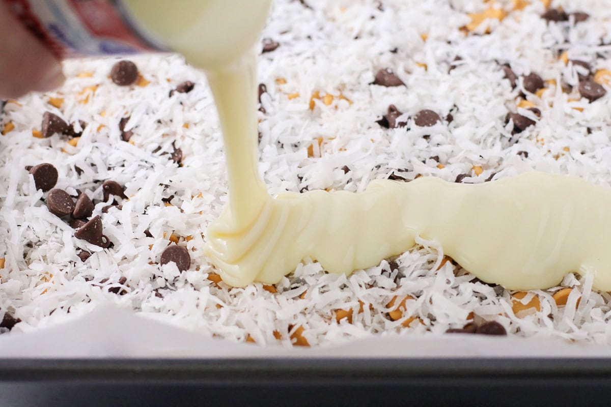 Pouring sweetened condensed milk on top of the layers of baking chips and coconut.
