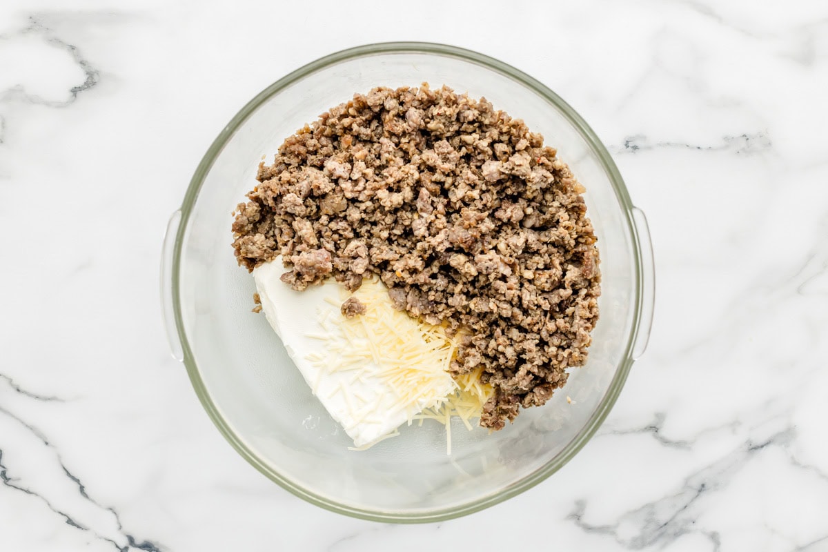 Combining cooked sausage crumbles, cream cheese, and shredded cheese in a glass bowl.