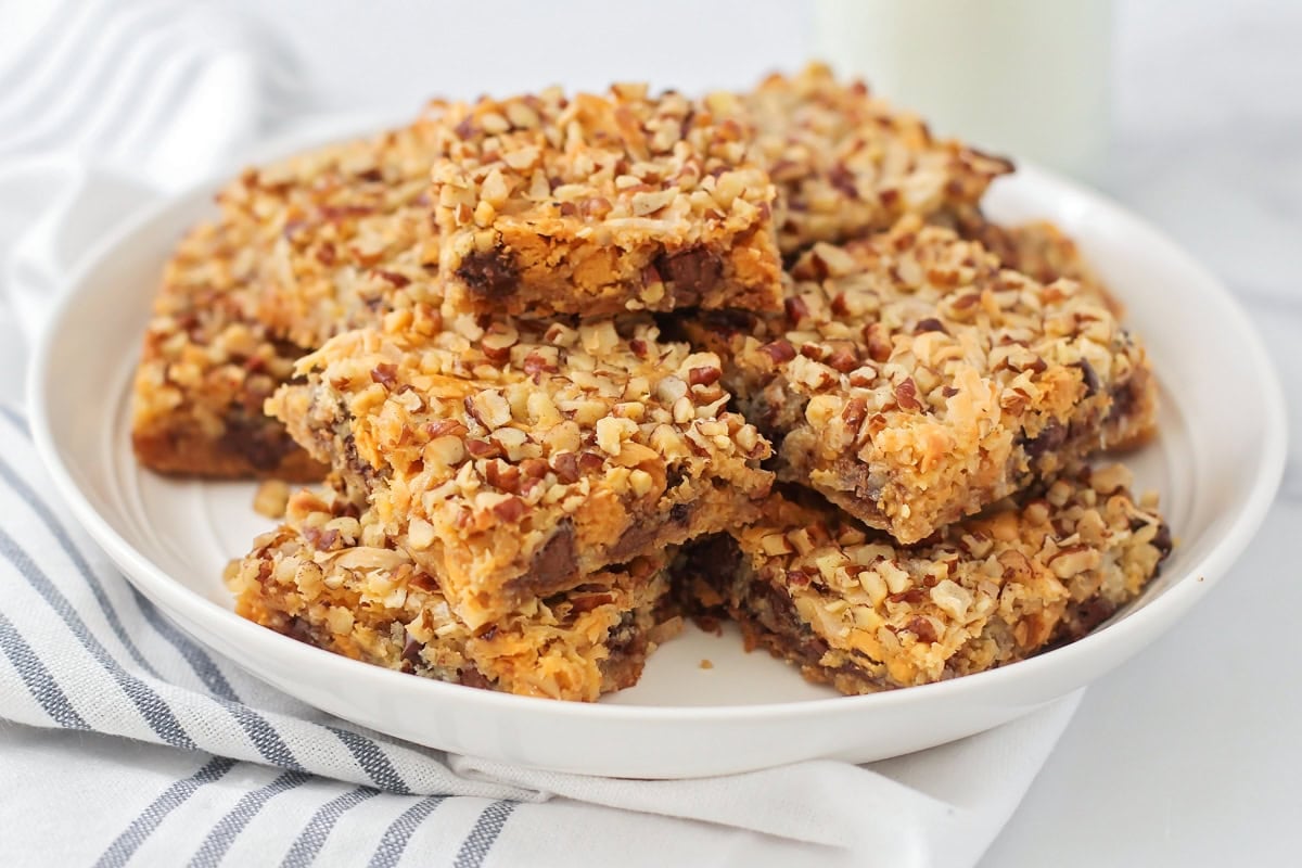 This homemade 7 layer bars recipe is rich, gooey, and loaded with chocolate, coconut, and nuts for the ultimate, easy dessert! 7 layer bars recipe squares stacked on a white plate.