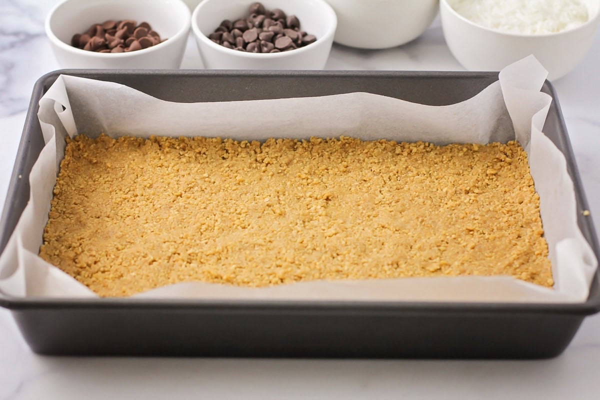 This homemade 7 layer bars recipe is rich, gooey, and loaded with chocolate, coconut, and nuts for the ultimate, easy dessert! Pressing a graham cracker crust into a baking dish.