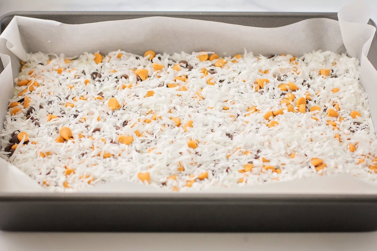 This homemade 7 layer bars recipe is rich, gooey, and loaded with chocolate, coconut, and nuts for the ultimate, easy dessert! Sprinkling coconut on top of the bars.