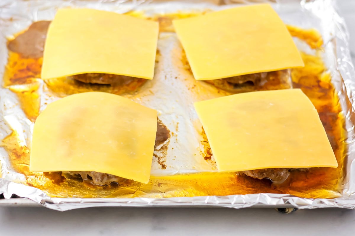 Burgers topped with cheese on a lined sheet pan.