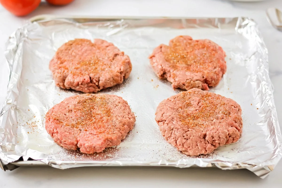 Shaped and seasoned burger patties on a lined sheet pan.