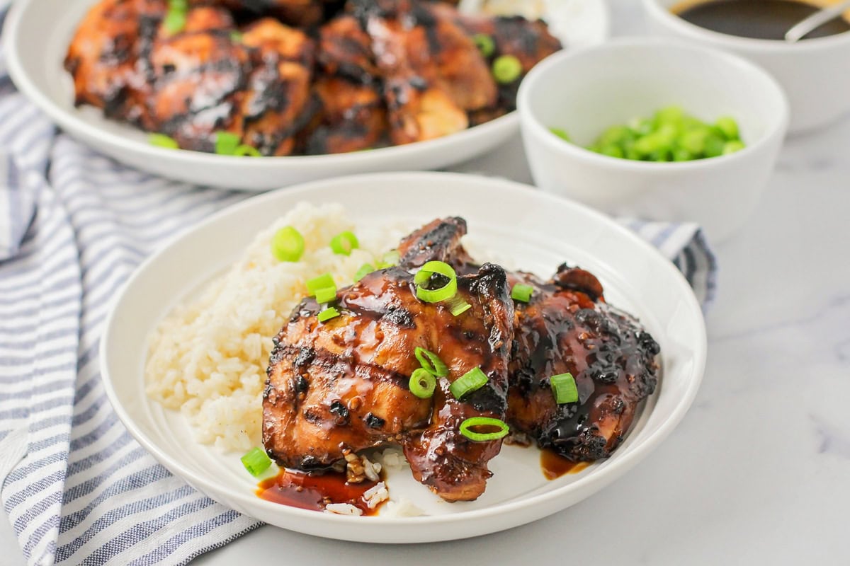 Grilled Huli Huli chicken is juicy, smoky, and glazed with a sweet and tangy Hawaiian-style marinade! Serve it alone or on rice or noodles. Grilled huli huli chicken served on a white plate with rice.