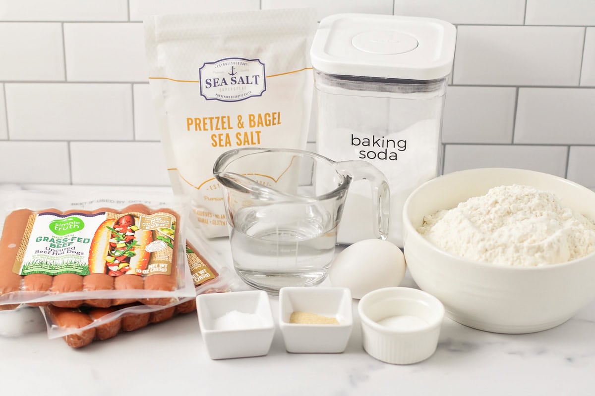 Our easy, homemade pretzel dogs will transport you to the mall, delivering that perfect chewy, salty pretzel wrapped around a savory hot dog! Pretzel dogs ingredients on a kitchen counter.