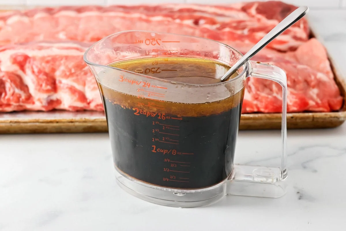 A measuring cup of marinade and a pan of ribs.