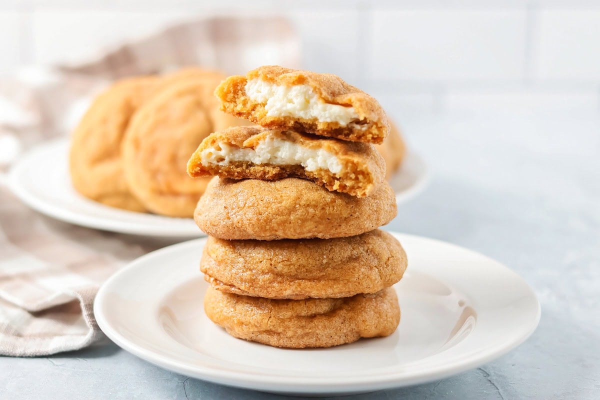 These delightful pumpkin cheesecake cookies combine two irresistible desserts into one irresistible bite, perfect for fall! Pumpkin cheesecake cookies stacked on each other on white plate.