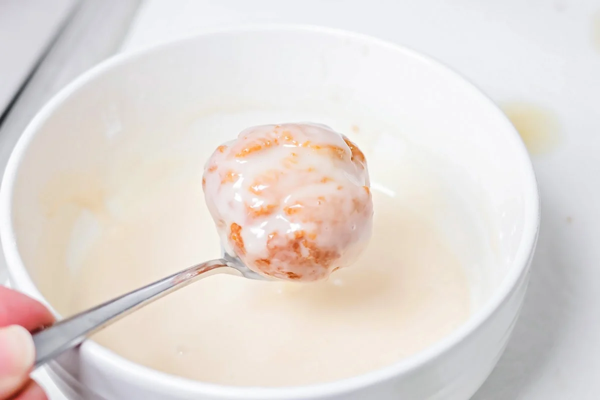A bonut coated with glaze on a spoon being held over a bowl of glaze.