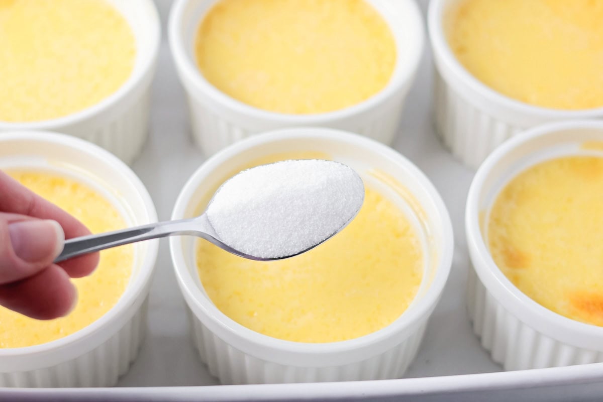 Holding a spoon of sugar over a baking dish with 6 baked creme brulees. 