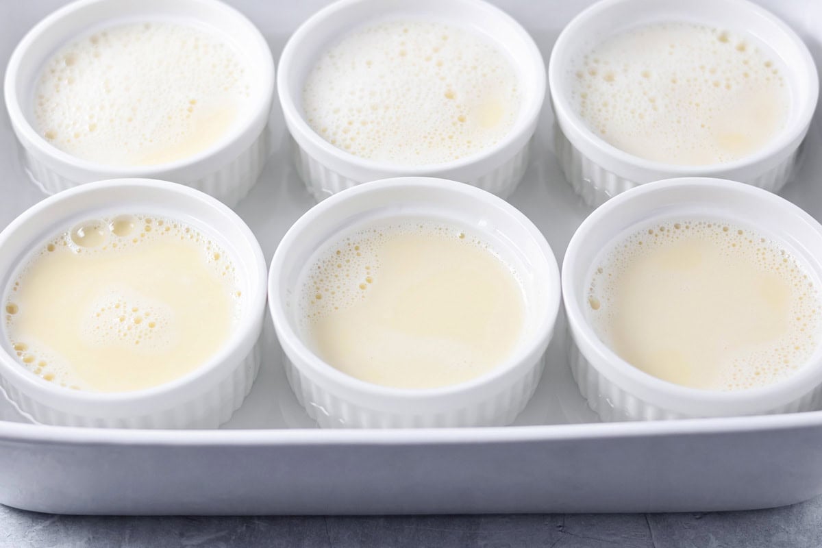 Six ramekins filled with a cream mixture in a baking dish with water added to reach halfway up the ramekins.