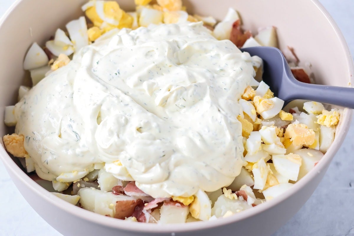 Creamy mayonnaise and dill mixture on top of diced red potatoes and hard boiled eggs in a mixing bowl.