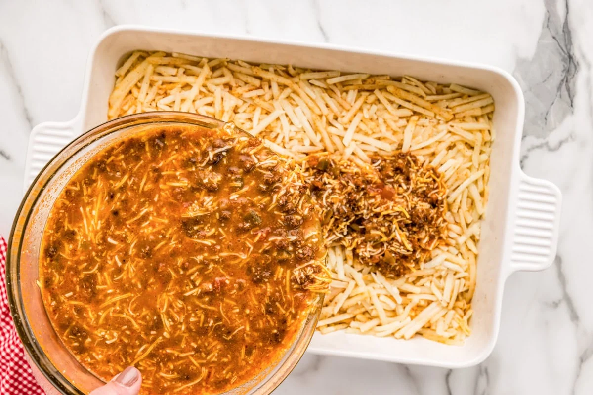 A bowl of savory custard mixed with cooked chorizo being poured over a layer of hash browns in a casserole dish.