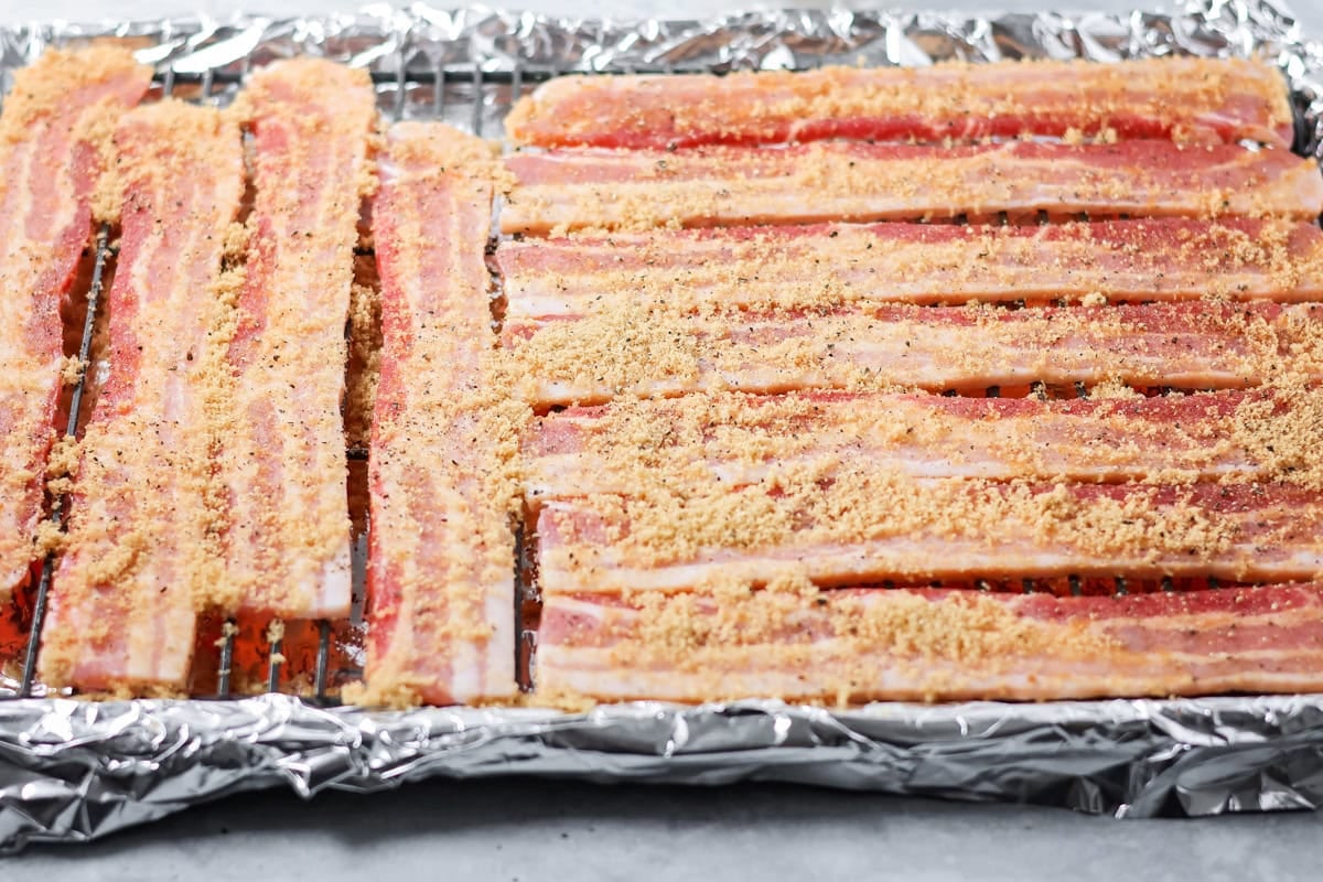 This 3-ingredient candied bacon is the perfect balance of sweet and salty and the ultimate upgrade to any gathering. Light brown sugar sprinkles over bacon strips on a wire racks.