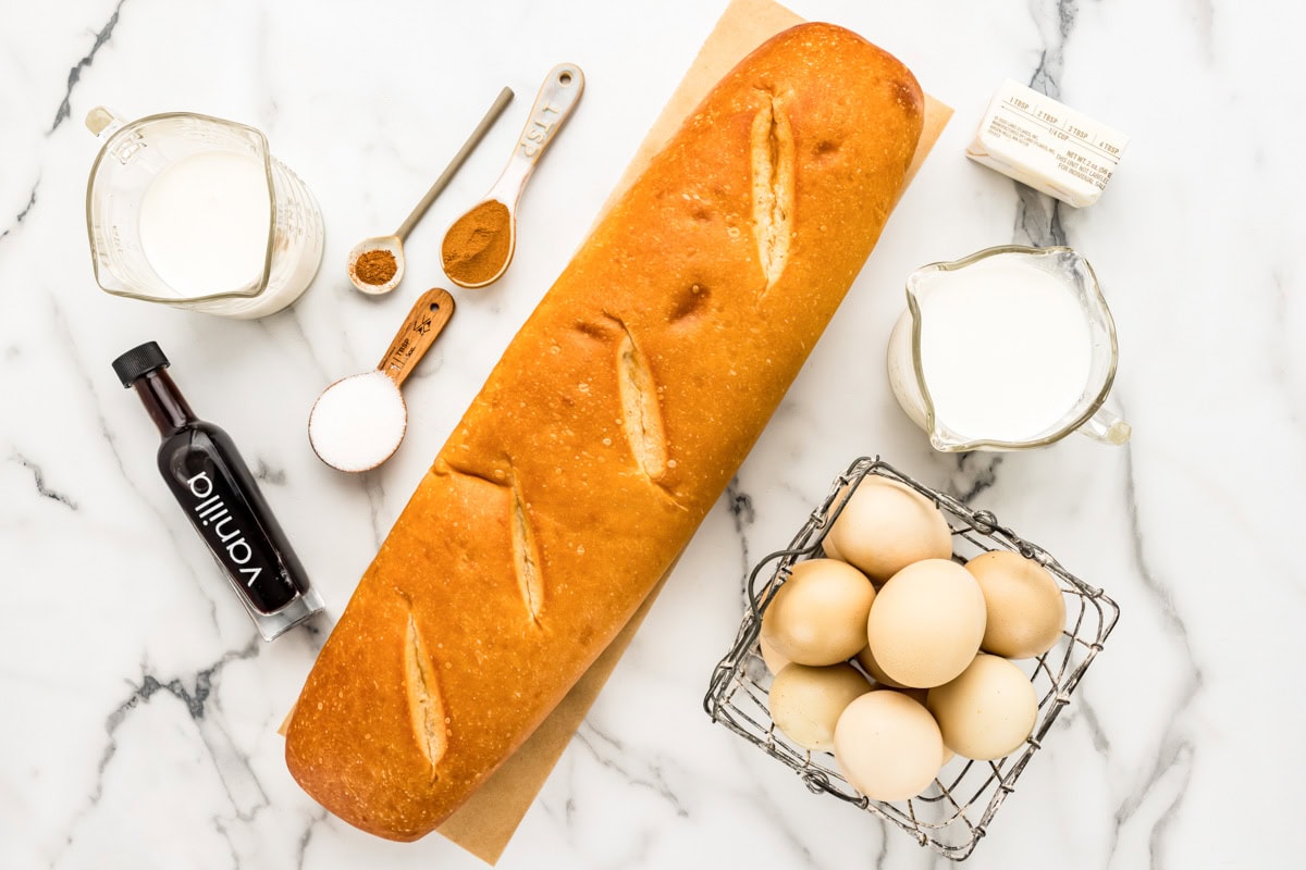 Sweet & filling oven French toast is perfectly spiced and can feed a crowd! This golden-baked goodness is a family favorite. Ingredients to make oven French toast on a kitchen counter.