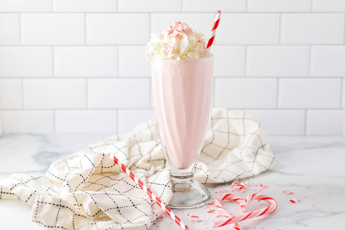 Celebrate the season with this festive peppermint milkshake - it's ready in minutes for a perfect holiday treat! Peppermint milkshake in a tall glass topped with whipped cream, peppermint candy sprinkles, and a red and white straw.