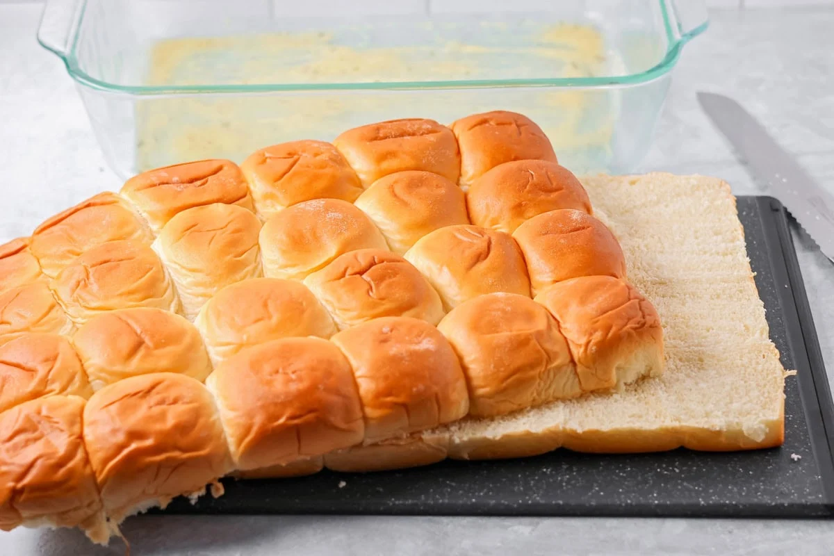 A block of 24 Hawaiin rolls sliced through the center next to a glass baking dish.