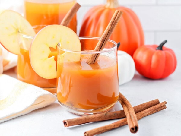 A glass of pumpkin juice with a cinnamon stick in it and apple slice.