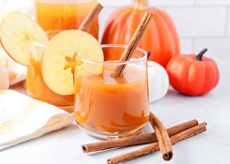 A glass of pumpkin juice with a cinnamon stick in it and apple slice.