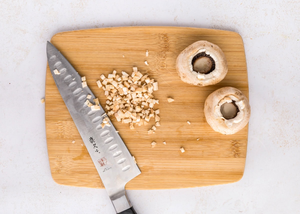 These cream cheese stuffed mushrooms are an easy, savory appetizer thatโs always the first to disappear at any party! Chopped mushrooms with a chefs knife on a wooden cutting board.
