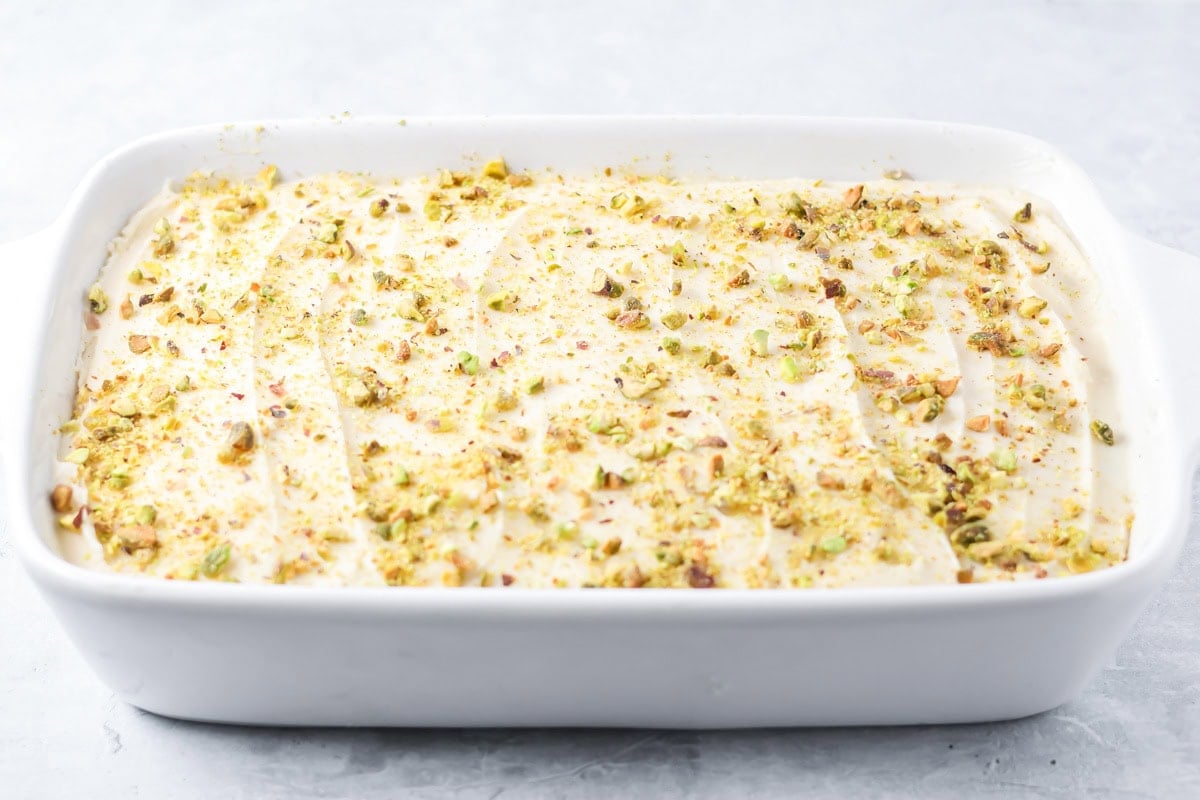 A baked pistachio cake topped with cream cheese icing and chopped pistachios on a kitchen counter.