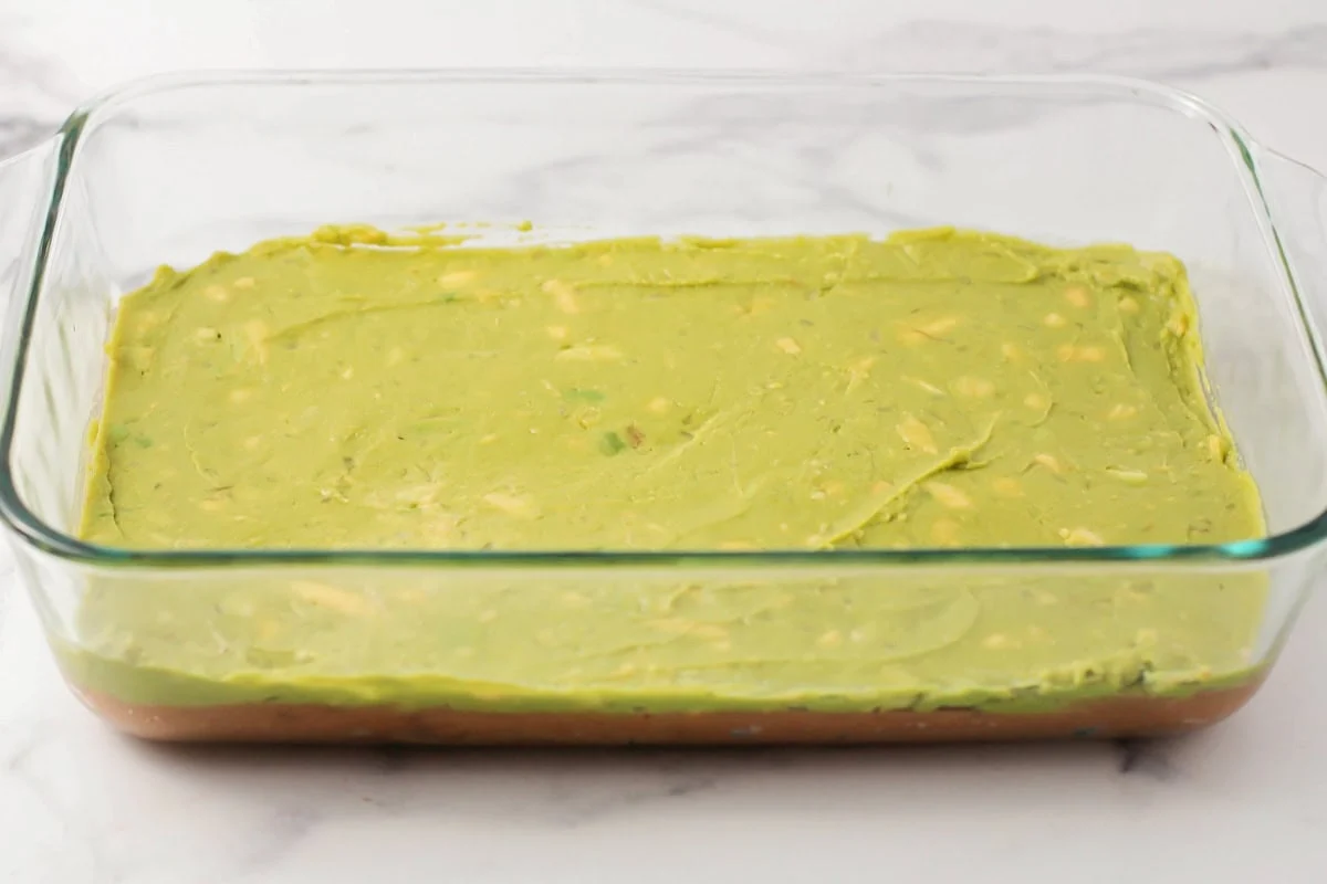 This 10-minute 7 layer bean dip is a must-have at every function! It has loads of flavor and all your favorite dip ingredients! A layer of guacamole spread in a baking dish.