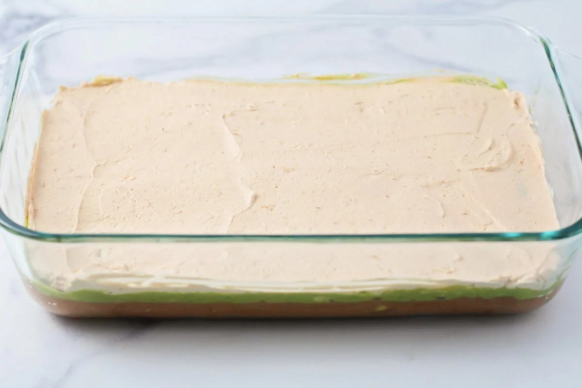 This 10-minute 7 layer bean dip is a must-have at every function! It has loads of flavor and all your favorite dip ingredients! A seasoned layer of sour cream spread in a baking dish.