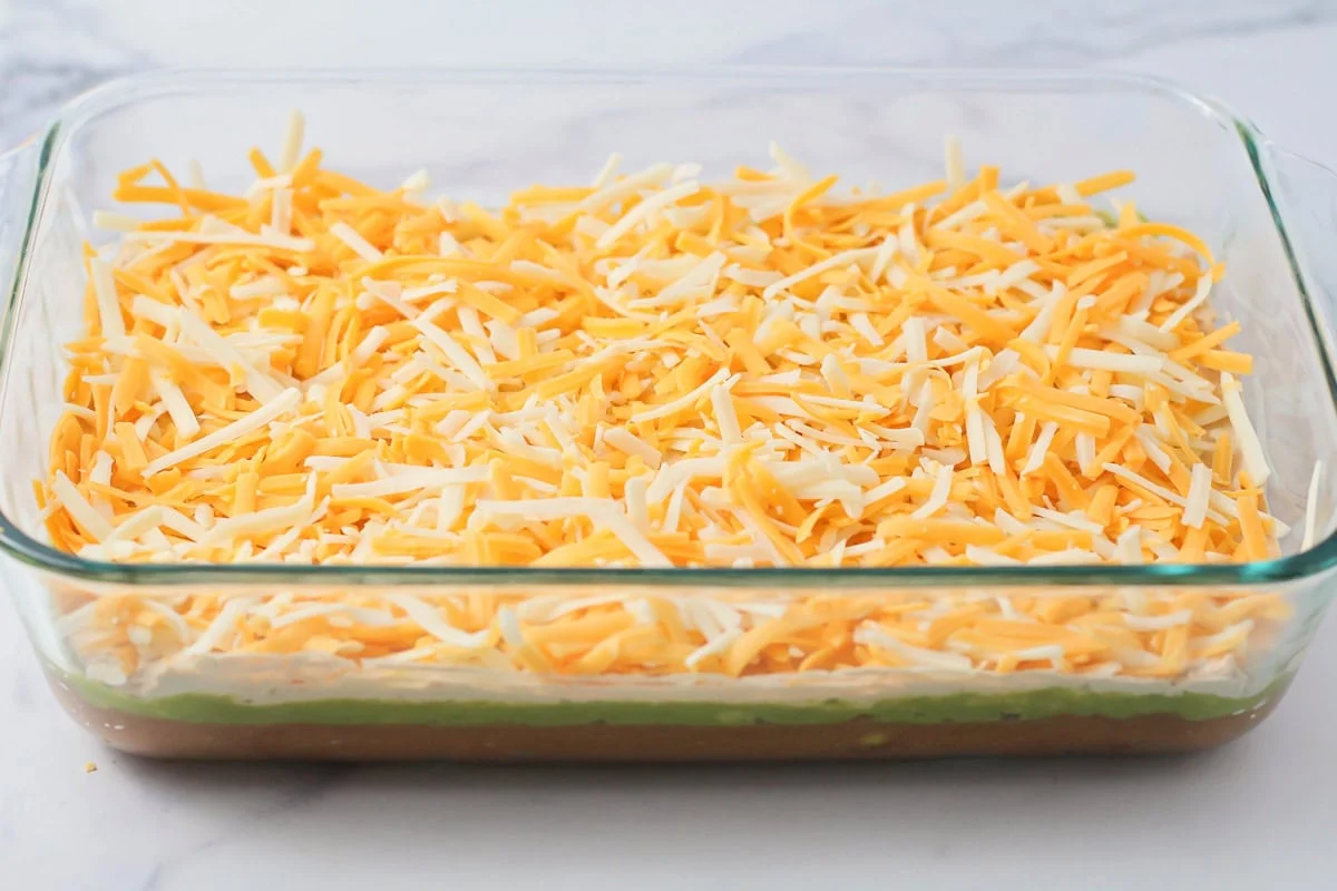 This 10-minute 7 layer bean dip is a must-have at every function! It has loads of flavor and all your favorite dip ingredients! A layer of shredded cheese in a baking dish.