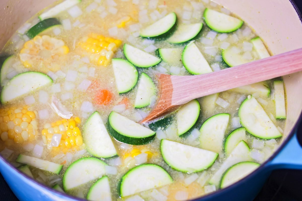 Grandma's famous caldo de pollo is a cozy chicken soup filled with chicken, rice, and Mexican spices and flavors! Chopped zucchini being added to chicken and corn cob soup.