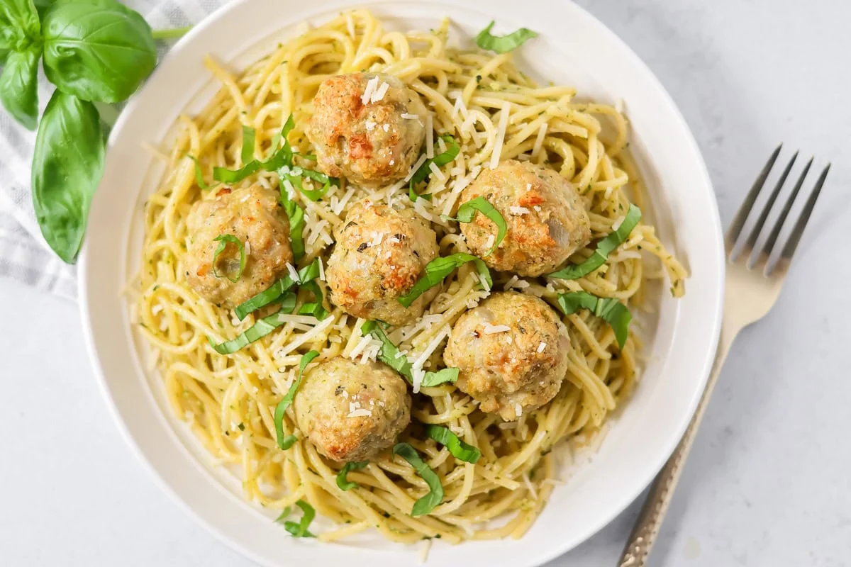 These easy chicken pesto meatballs come together in 30 minutes for a flavorful, family-friendly dinner full of basil and parmesan goodness. Chicken pesto meatballs on a bed of pasta on a serving plate.