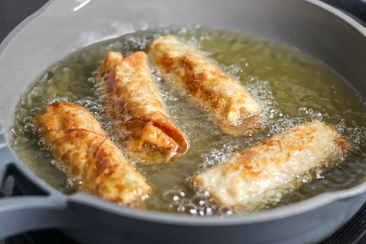 Four egg rolls frying in a skillet of oil.