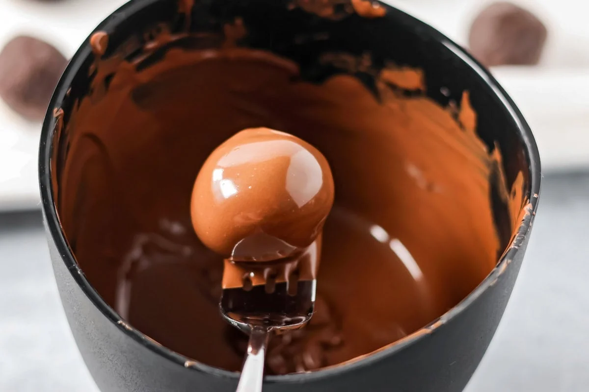 No-bake Oreo cream cheese balls are a three ingredient treat with a silky center and chocolate coating, ideal for every occasion. Holding chocolate ball dipped in chocolate with a fork over a bowl of melted chocolate.