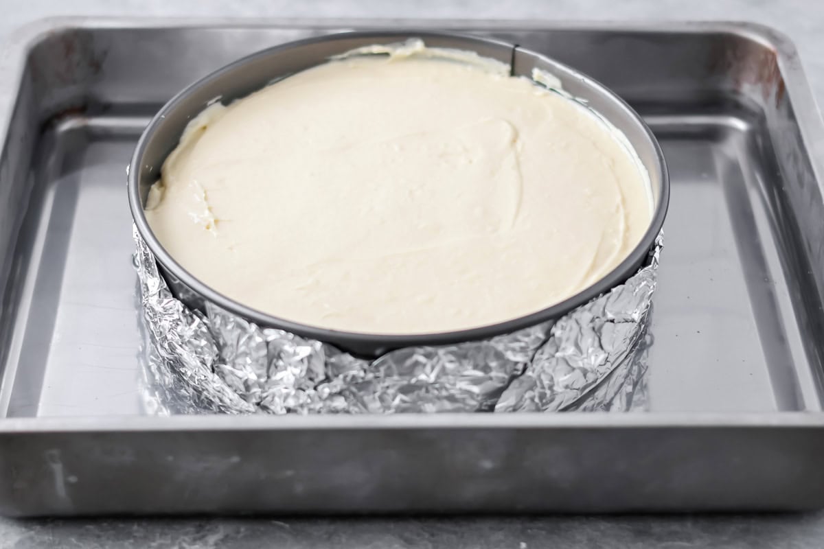 Plain cheesecake filling spread on top of caramel sauce. The aluminum foil wrapped springform pan is in a pan with water. 