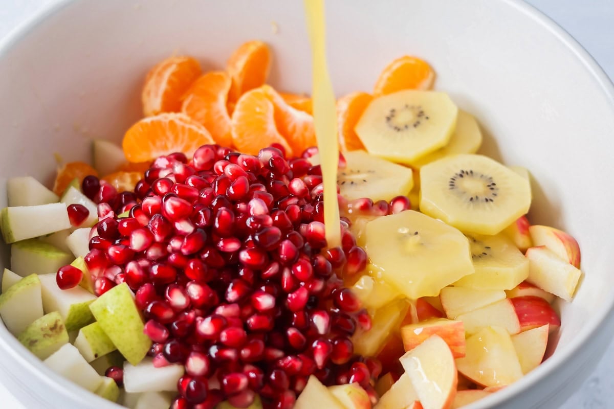 Bright and refreshing, this winter fruit salad blends clementines, kiwi, apples, pear, and pomegranate with a light citrus honey dressing. Pouring citrus honey dressing over the fruit in the bowl.