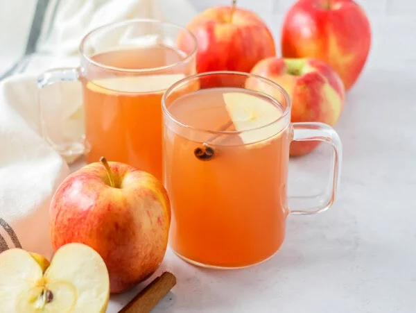 Homemade apple cider poured into 2 cups with apples and cinnamon sticks around.
