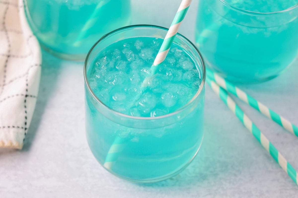 Blue punch recipe in glass with ice and straw.