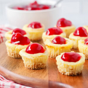 Mini cheesecake bites on serving board with cherries in the background.