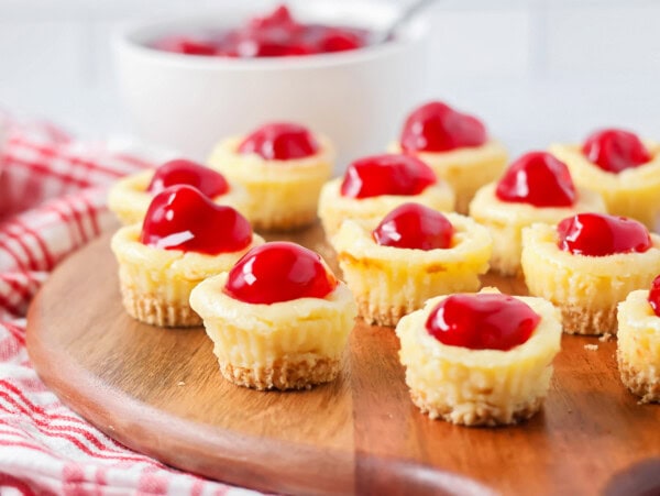 Mini cheesecake bites on serving board with cherries in the background.