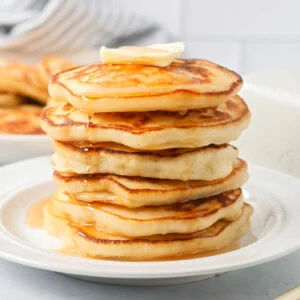 Easy pancake recipe stacked on top of each other on white plate with butter and syrup.
