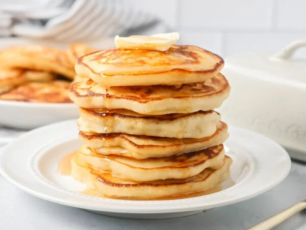 Easy pancake recipe stacked on top of each other on white plate with butter and syrup.
