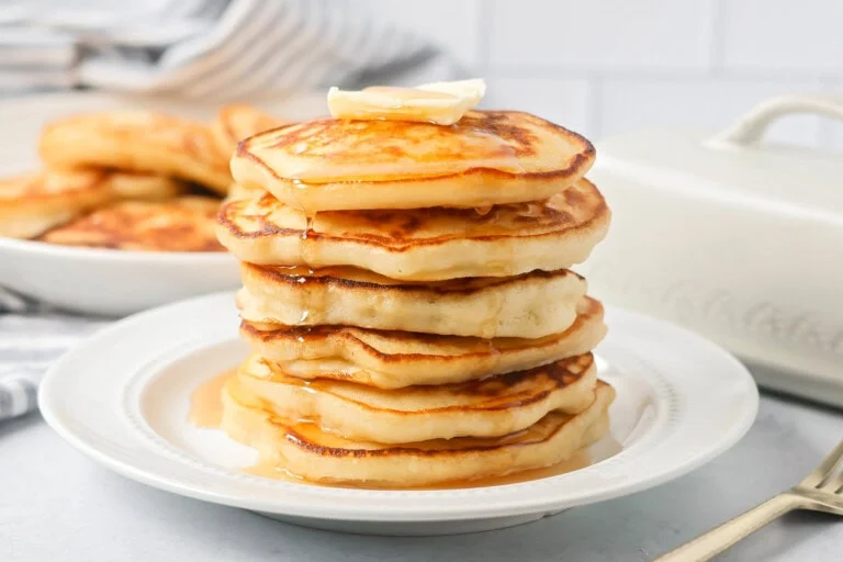 Easy pancake recipe stacked on top of each other on white plate with butter and syrup.