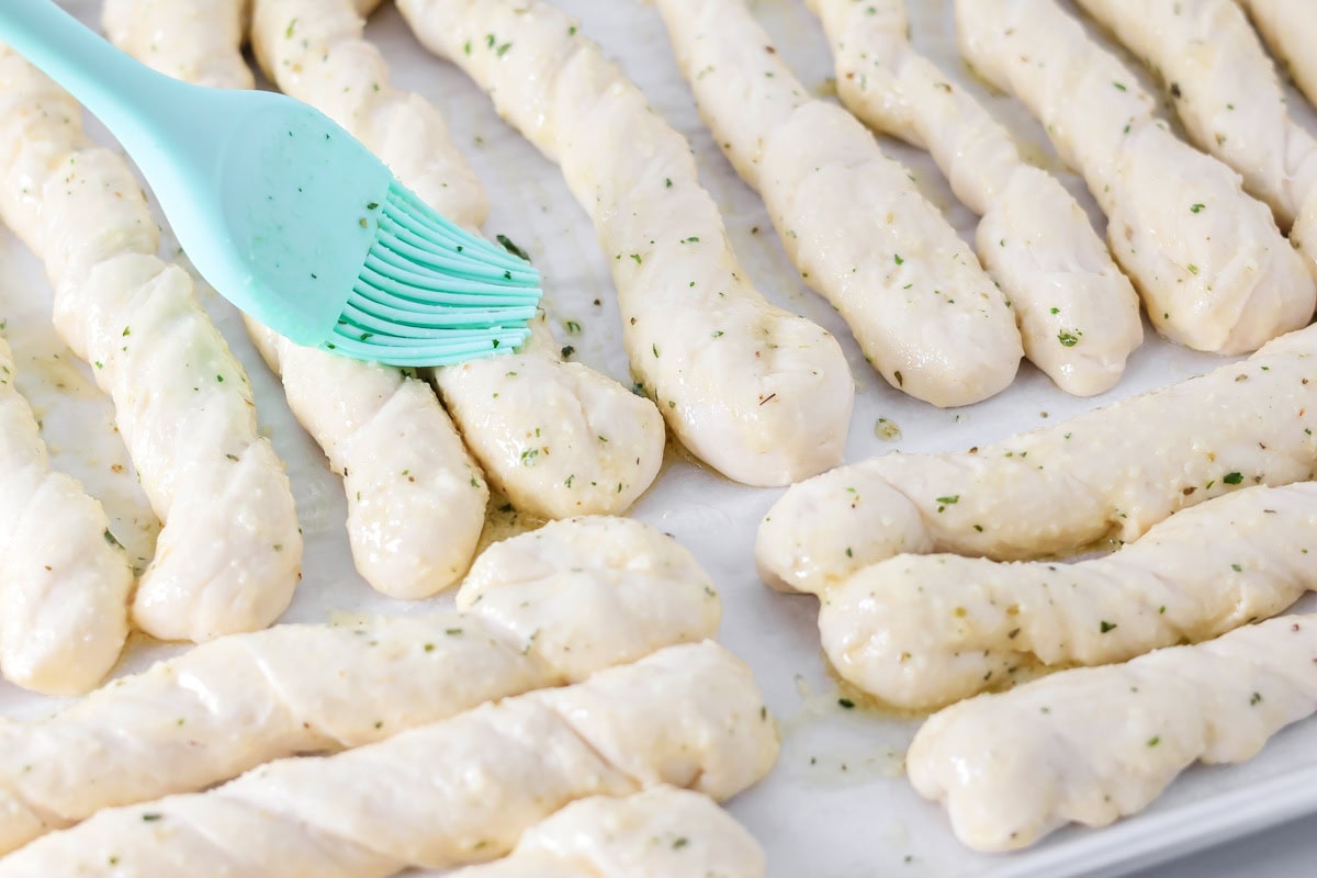 Brushing breadsticks with seasonings on a lined baking sheet.