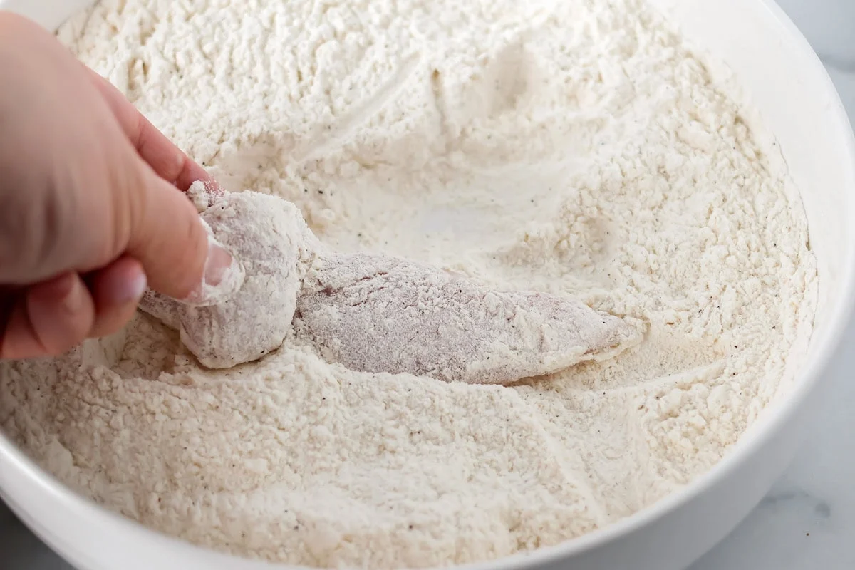 Dipping buffalo chicken tenders into a bowl of flour.